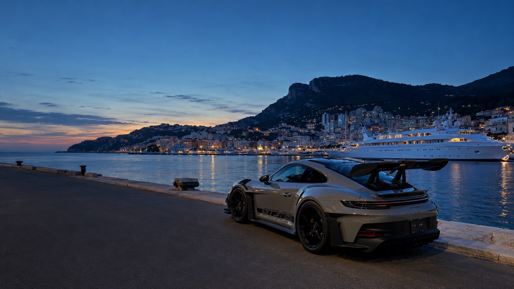 A grey Porsche 911 GT3 RS parked on a harbor quay at dusk beside a superyacht, with a lit hillside town behind